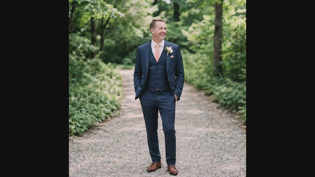 A smiling man walks forward along a gravel path in a wooded area, looking off to the side. The camera zooms in on his torso and face as he walks, showcasing his three-piece navy blue suit, white shirt, peach necktie, peach pocket square, and boutonniere.